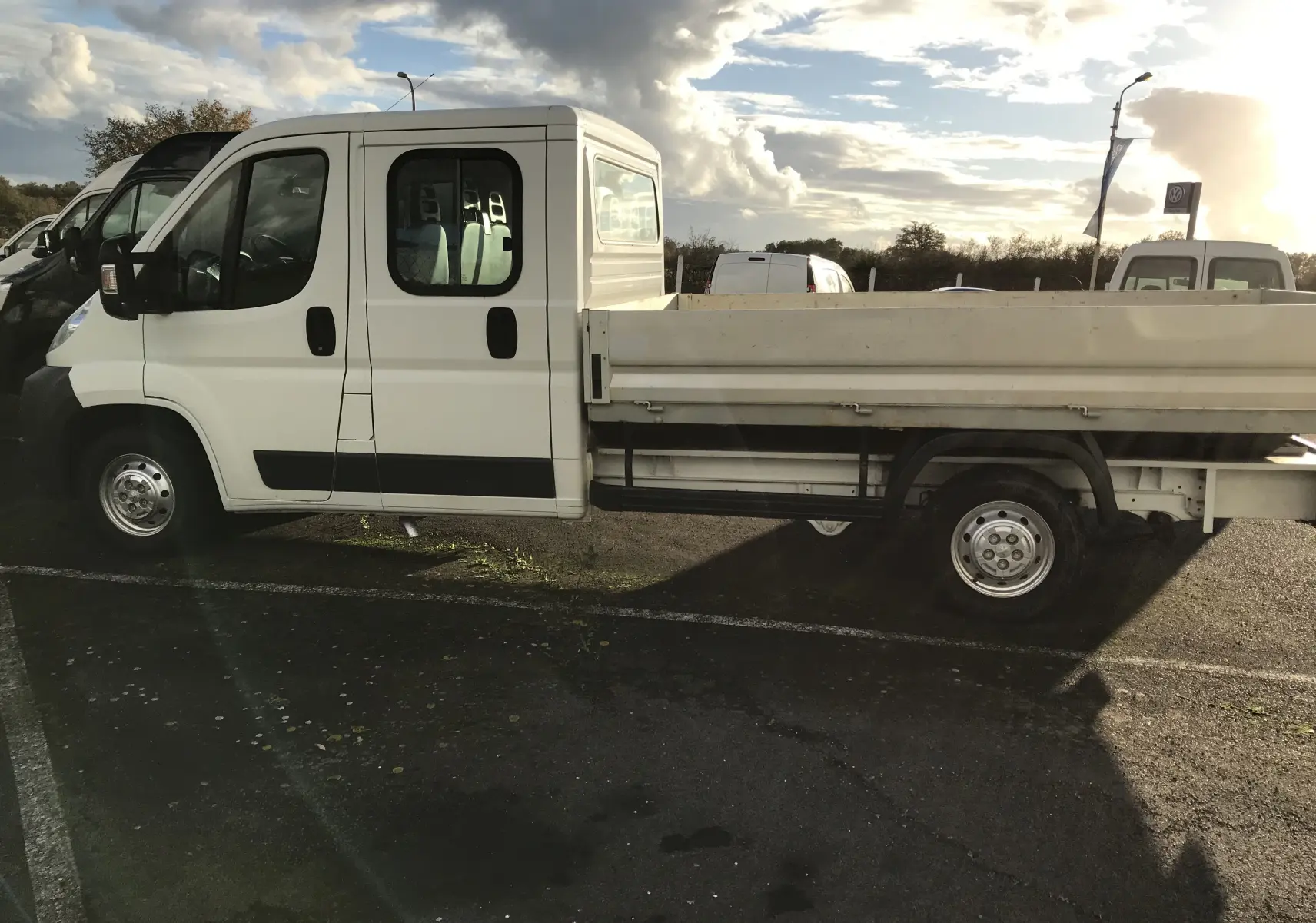 Vue latérale côté droit d’un Peugeot Boxer Châssis Double Cabine blanc avec plateau, stationné en extérieur sous un ciel nuageux.