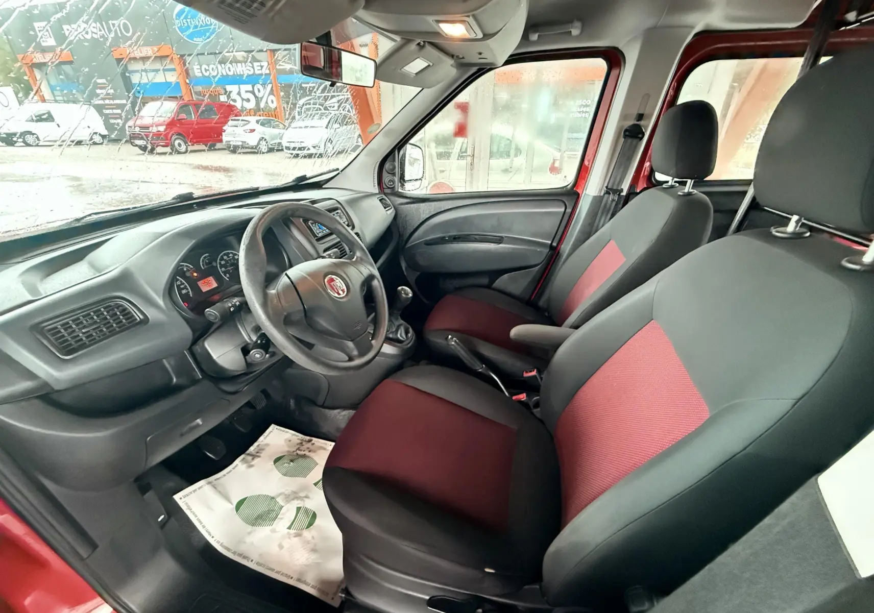 Intérieur du Fiat Doblo Cargo 2011 vu côté conducteur, sièges tissu rouge et noir, volant cuir et tableau de bord noir.
