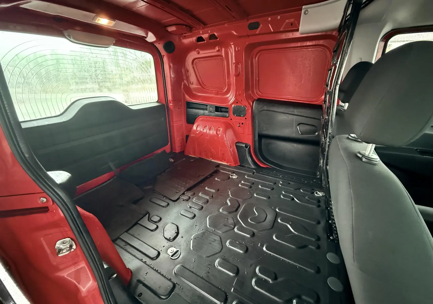 Vue intérieure arrière du Fiat Doblo Cargo rouge de 2011, montrant l’espace de chargement vide avec plancher noir et sièges avant partiellement visibles.