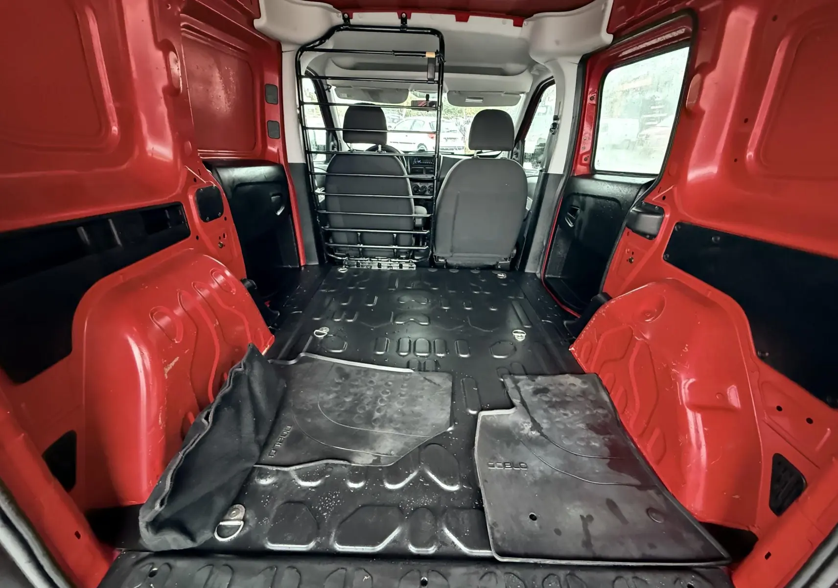 Vue intérieure arrière du FIAT Doblo Cargo rouge, montrant l'espace de chargement vide avec tapis noirs et séparation métallique.