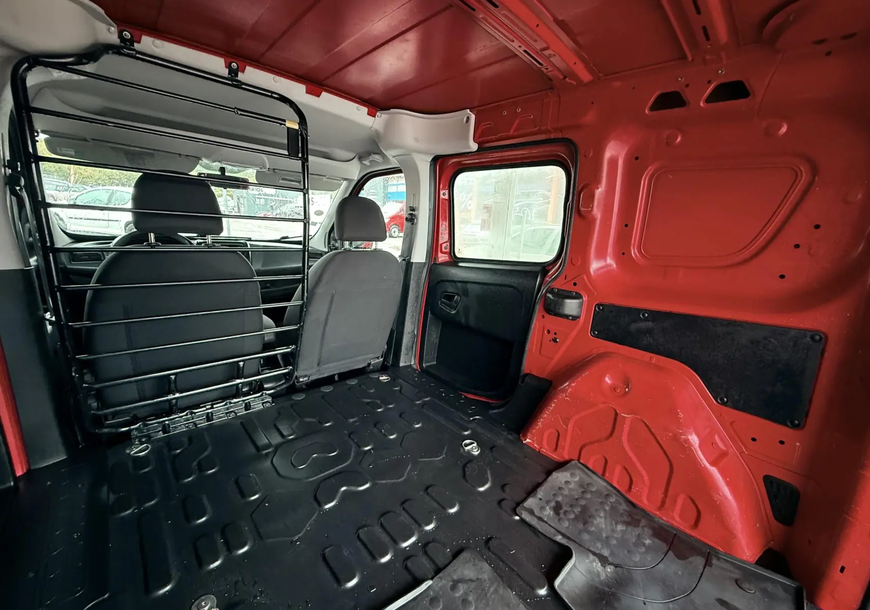 Vue intérieure arrière du Fiat Doblo Cargo rouge, montrant l'espace de chargement vide et la séparation métallique avec les sièges avant.