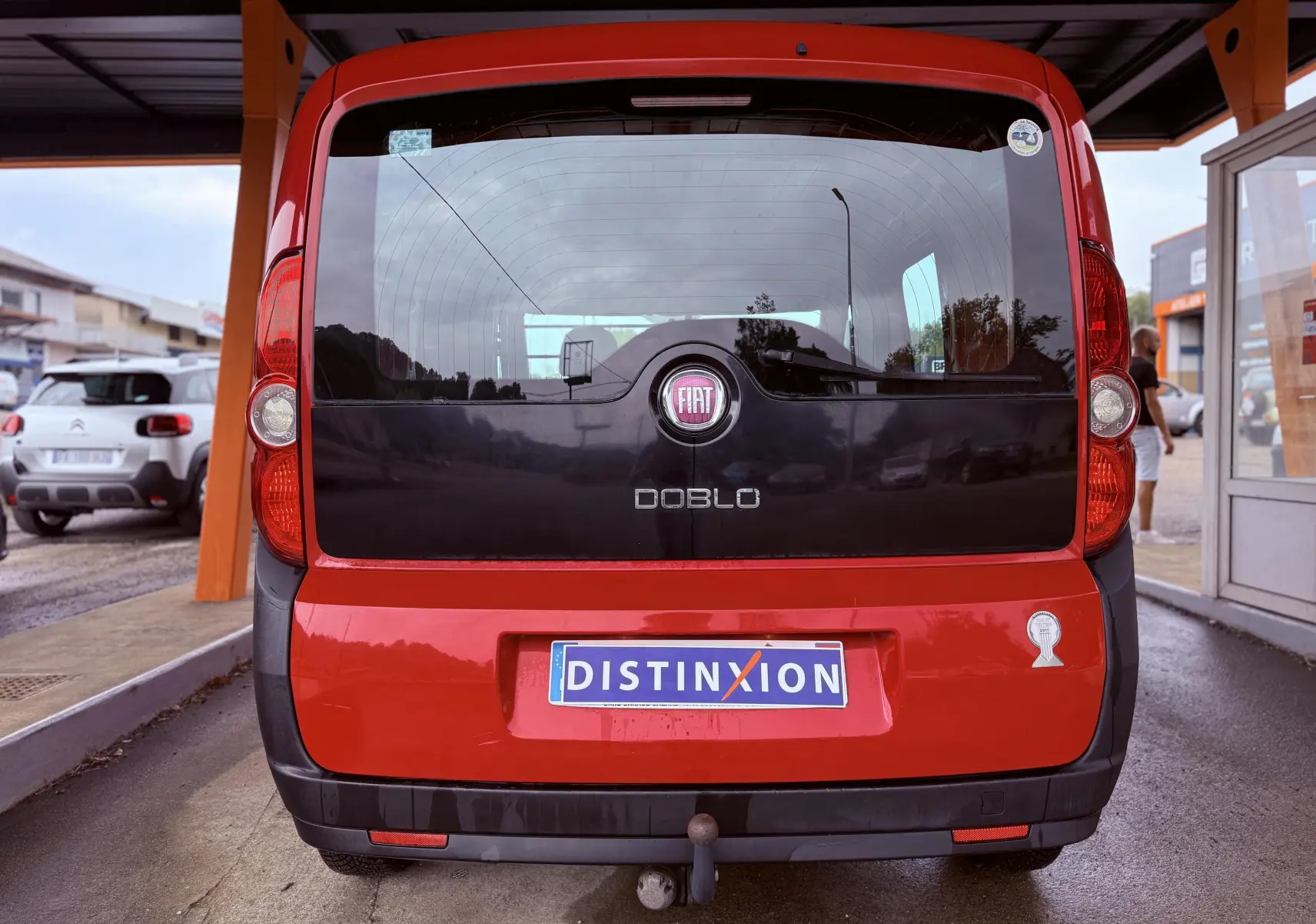 Vue arrière d'un Fiat Doblo Cargo rouge avec hayon noir et attelage sous un abri.