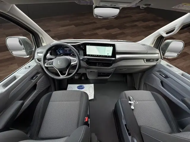 Vue intérieure avant du Volkswagen Transporter ProCab 2026 gris, avec tableau de bord moderne, volant multifonction et sièges tissu rayé.