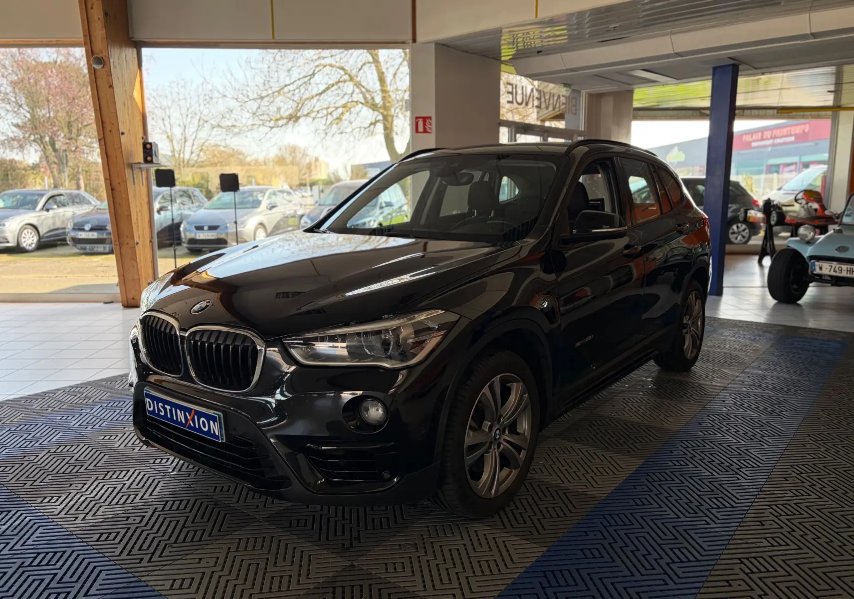 BMW X1 noir en 3/4 avant droit, exposée en intérieur avec calandre et phares LED bien visibles.