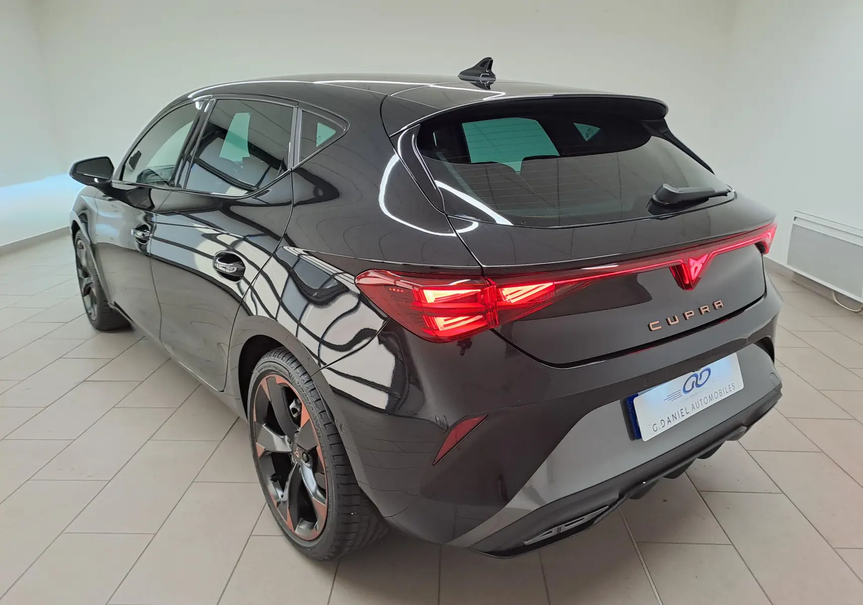 Vue 3/4 arrière droite d'une CUPRA Leon Ph.2 noire Minuit avec jantes cuivrées et feux arrière allumés dans un showroom.