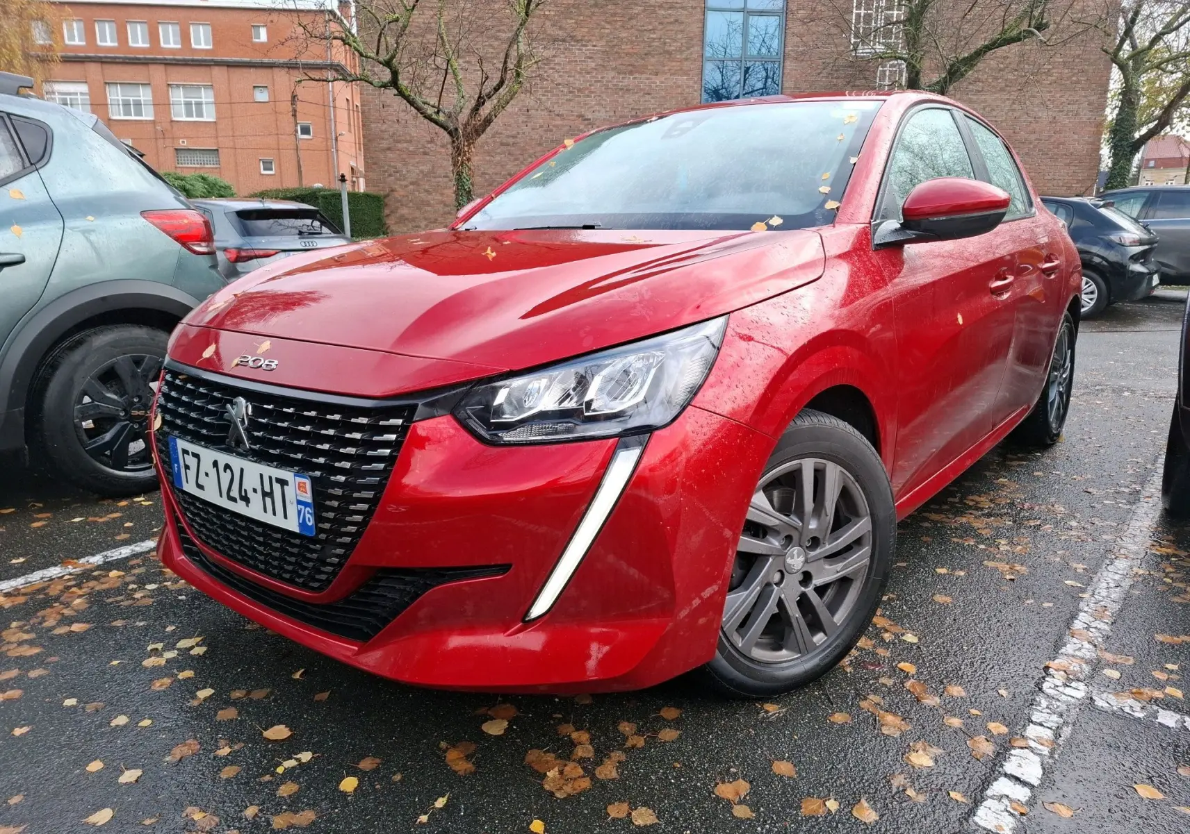 Peugeot 208 rouge Elixir vue en 3/4 avant droit, avec calandre noire et feux LED distinctifs sous la pluie.