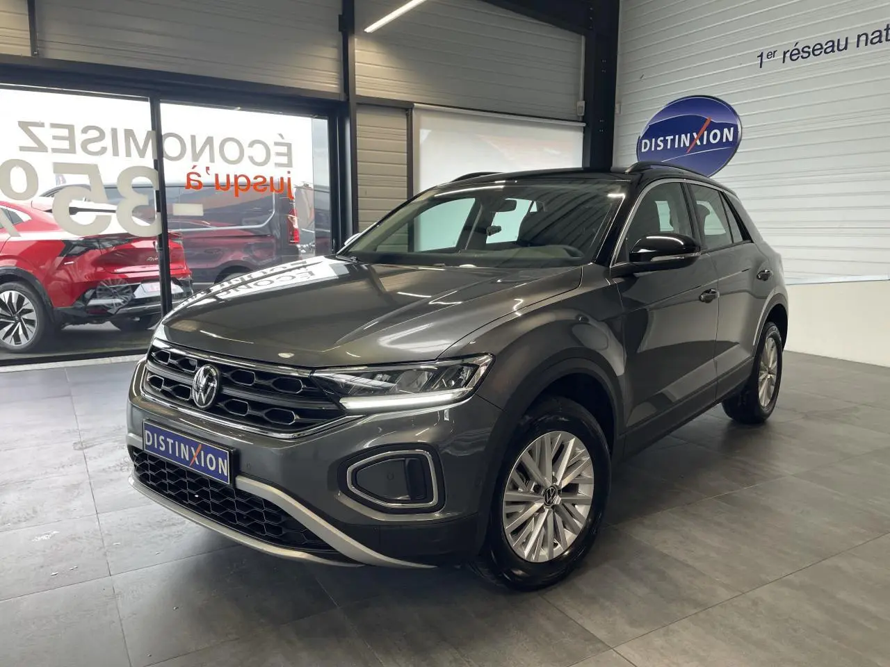 Volkswagen T-Roc 2025 gris Indium en 3/4 avant droit avec toit noir et jantes alliage 16 pouces dans un showroom.