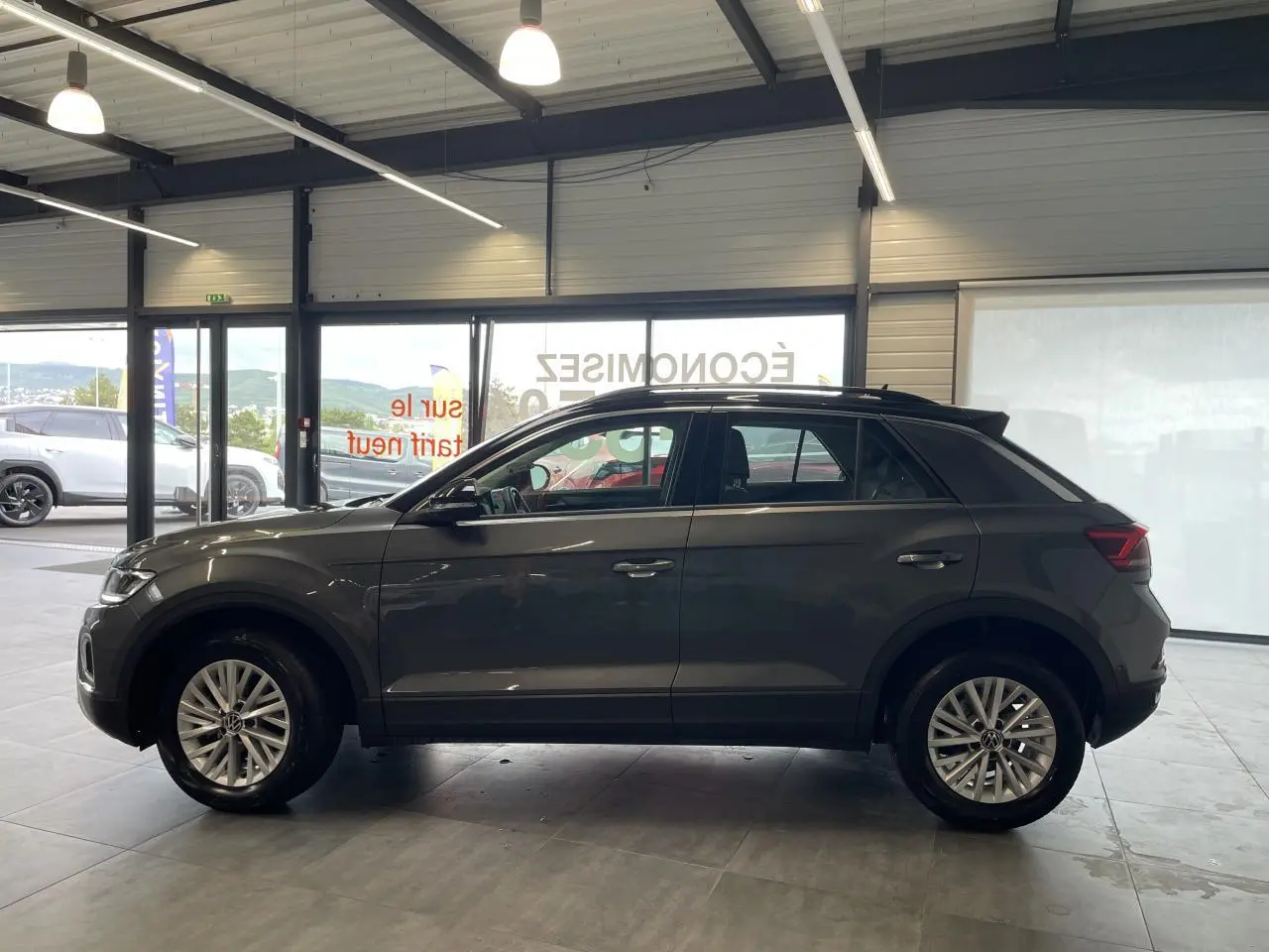 Volkswagen T-Roc 2025 gris Indium, vue de profil côté gauche en intérieur avec jantes alliage et toit noir.