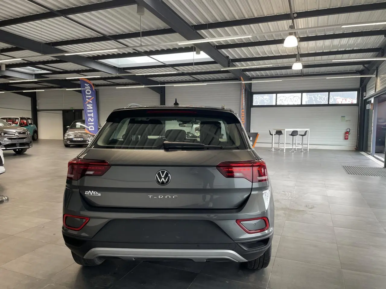 Vue arrière du Volkswagen T-Roc 2025 en gris Indium métallisé avec feux LED et diffuseur argenté dans un showroom.