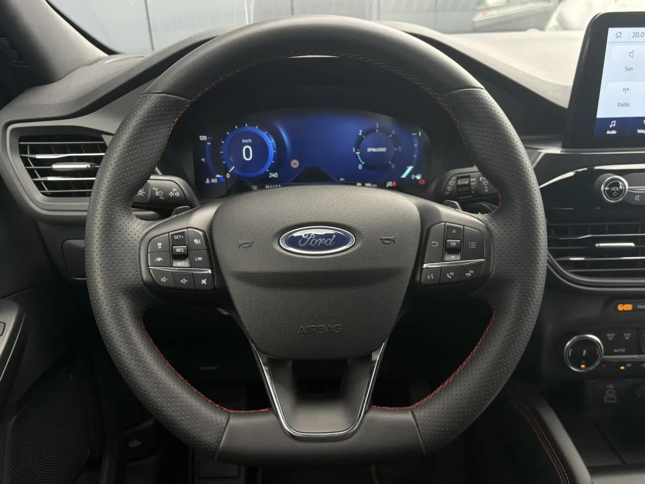 Vue rapprochée du volant noir perforé avec surpiqûres rouges du Ford Kuga 2023, tableau de bord numérique en arrière-plan.