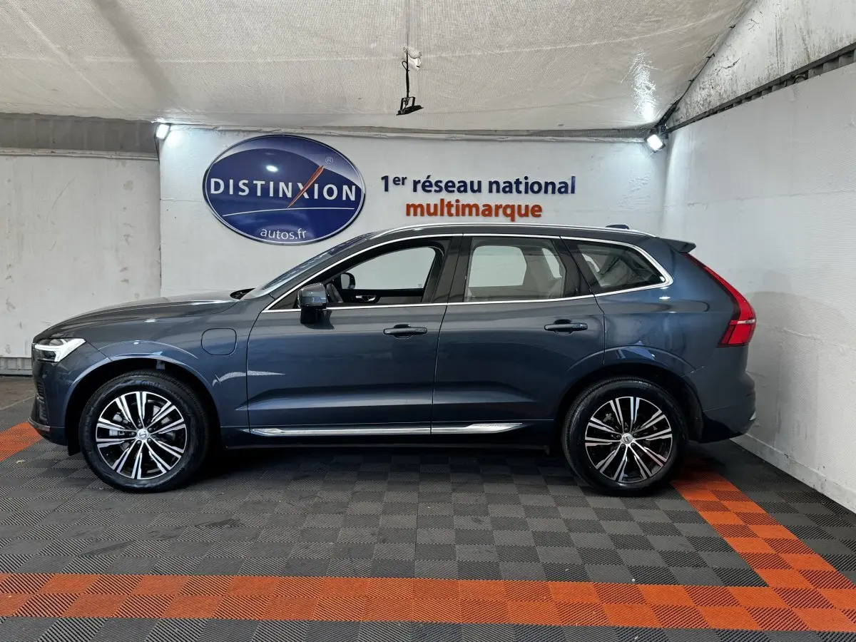 Profil droit d’un Volvo XC60 bleu hybride rechargeable avec jantes alliage et rails de toit chromés, en intérieur showroom.
