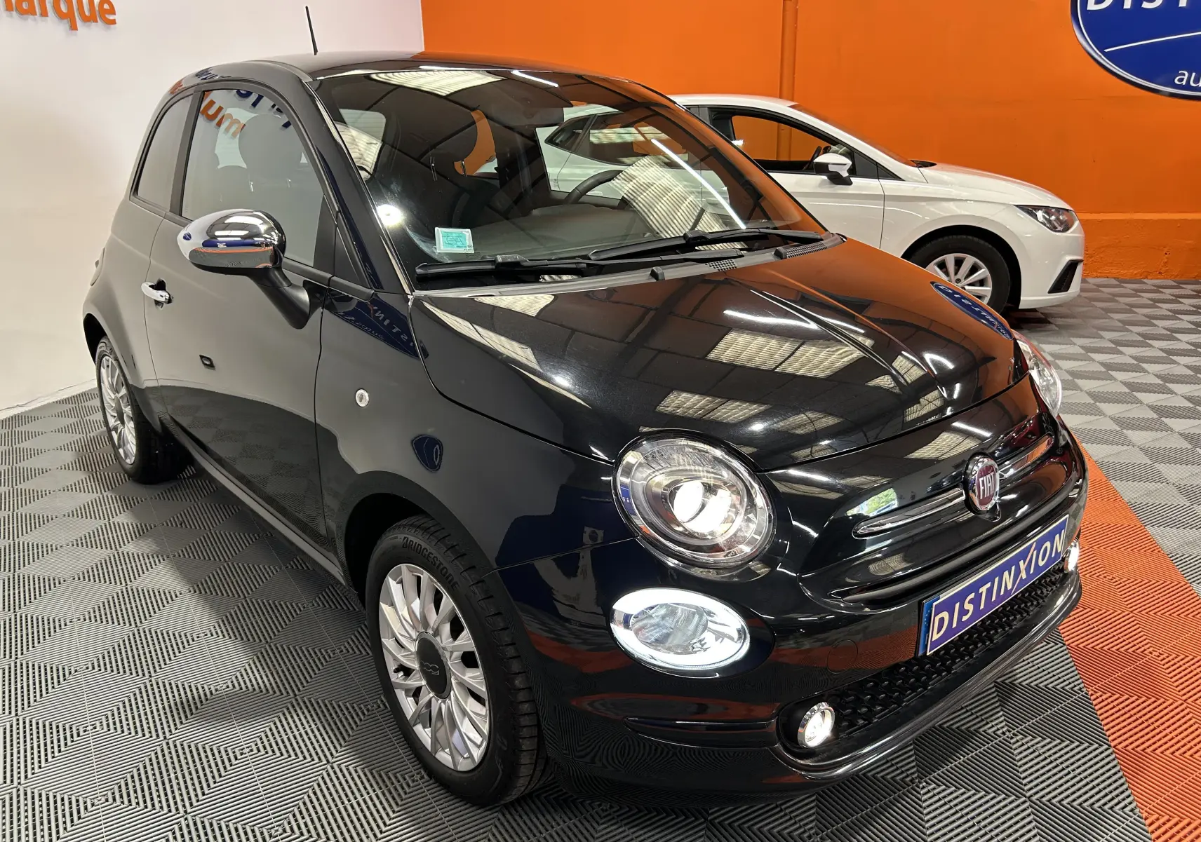 Fiat 500 noire vue 3/4 avant droit dans un showroom avec phares et antibrouillards allumés.
