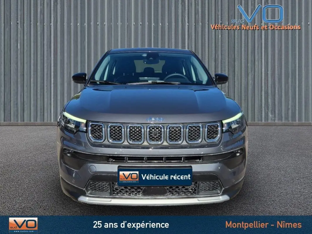 Vue frontale d'un Jeep Compass gris 2024 avec calandre distinctive et phares LED allumés sur fond industriel.