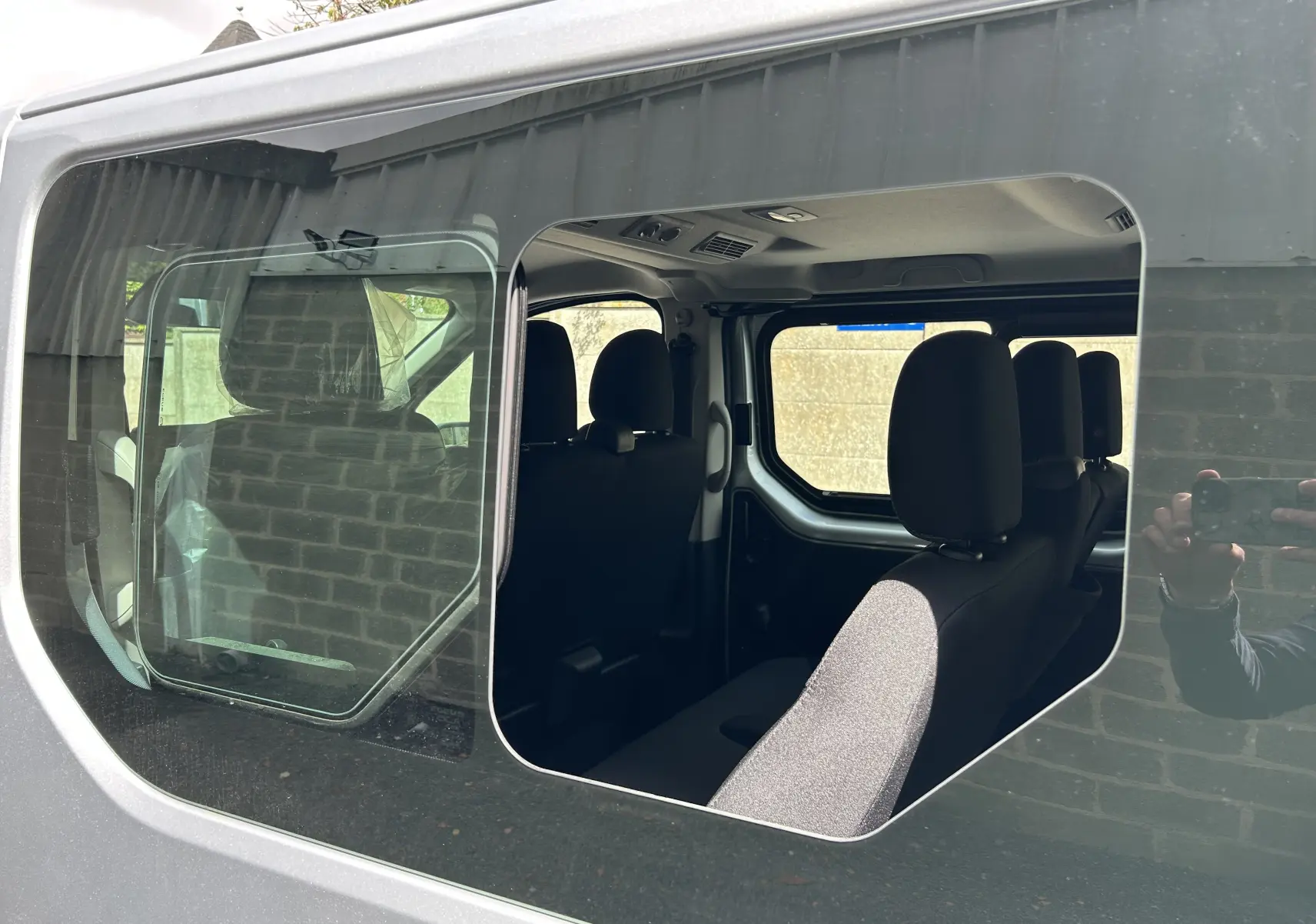 Vue intérieure par la porte latérale gauche ouverte du Renault Trafic Combi blanc glacier, montrant les sièges en tissu noir.