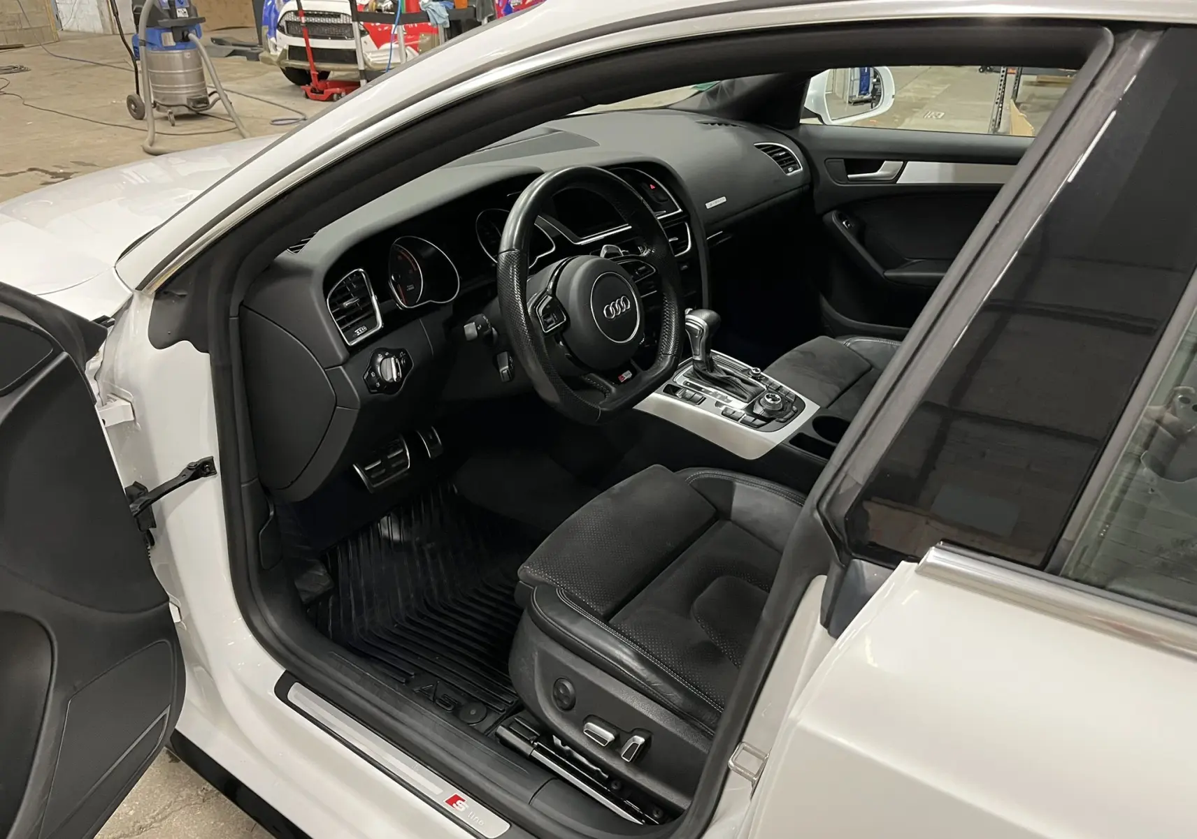 Vue intérieure côté conducteur d’une Audi A5 Sportback blanche de 2015, avec sellerie cuir noir et tableau de bord moderne.