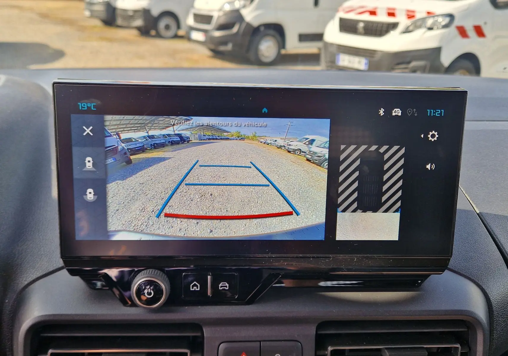 Écran central du Citroën Berlingo gris Artense 2025 montrant la caméra de recul avec vue arrière sur un parking de véhicules blancs.