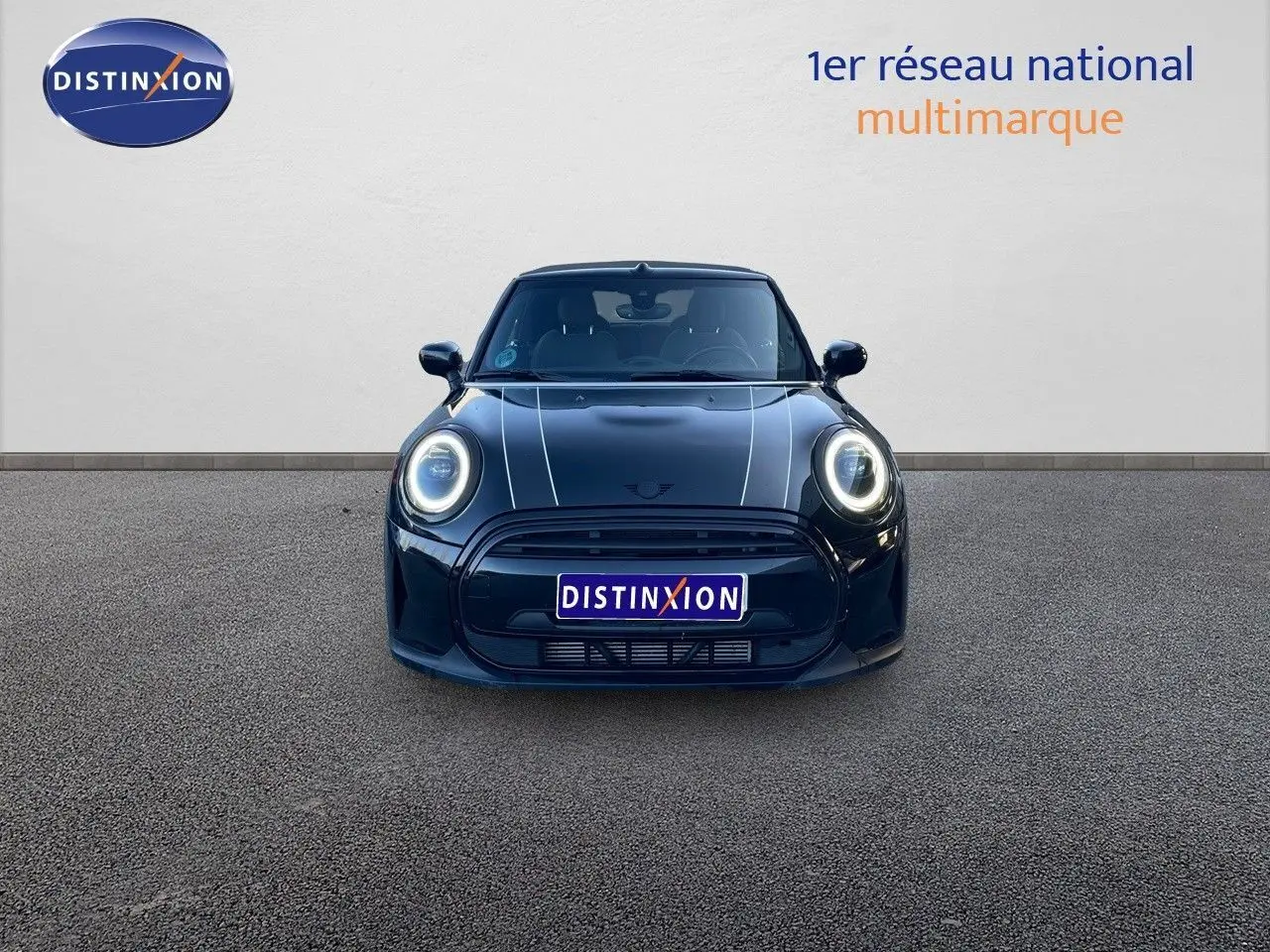 Vue frontale d'une MINI Cabriolet F57 noir midnight metal avec phares LED allumés et capot à bandes blanches.