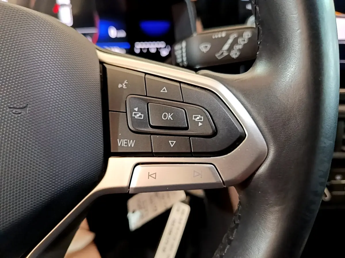 Gros plan sur les commandes gauche du volant cuir alu du Volkswagen Taigo noir, avec boutons multifonctions et affichage numérique flou.