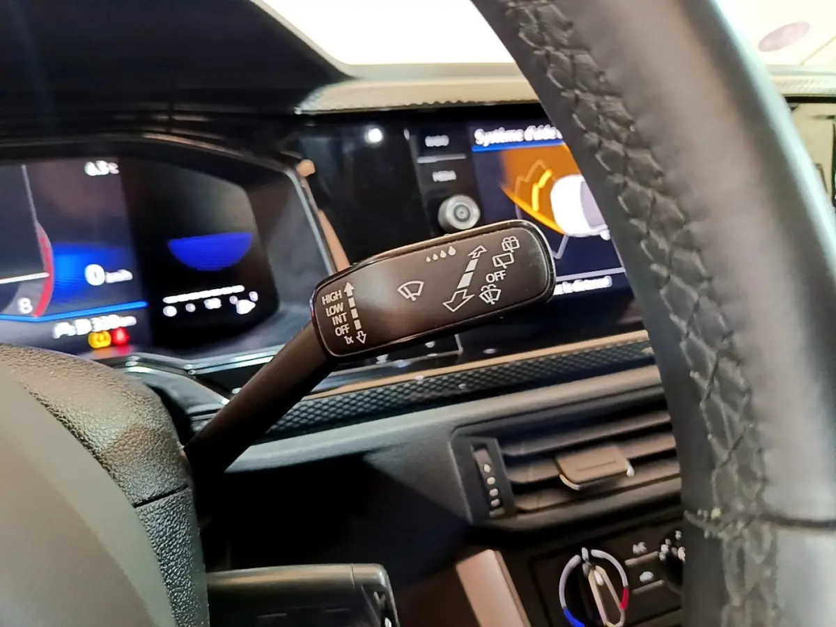 Gros plan sur la commande d’essuie-glace à gauche du volant cuir du Volkswagen Taigo noir, tableau de bord numérique en arrière-plan.