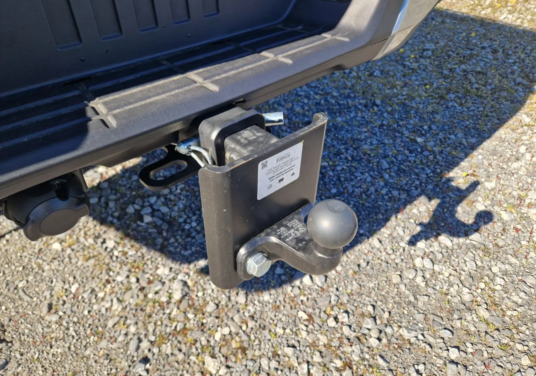 Gros plan sur l'attelage arrière noir du Ford Ranger Super Cabine gris Iconic, avec prise électrique visible, sur sol gravillonné.