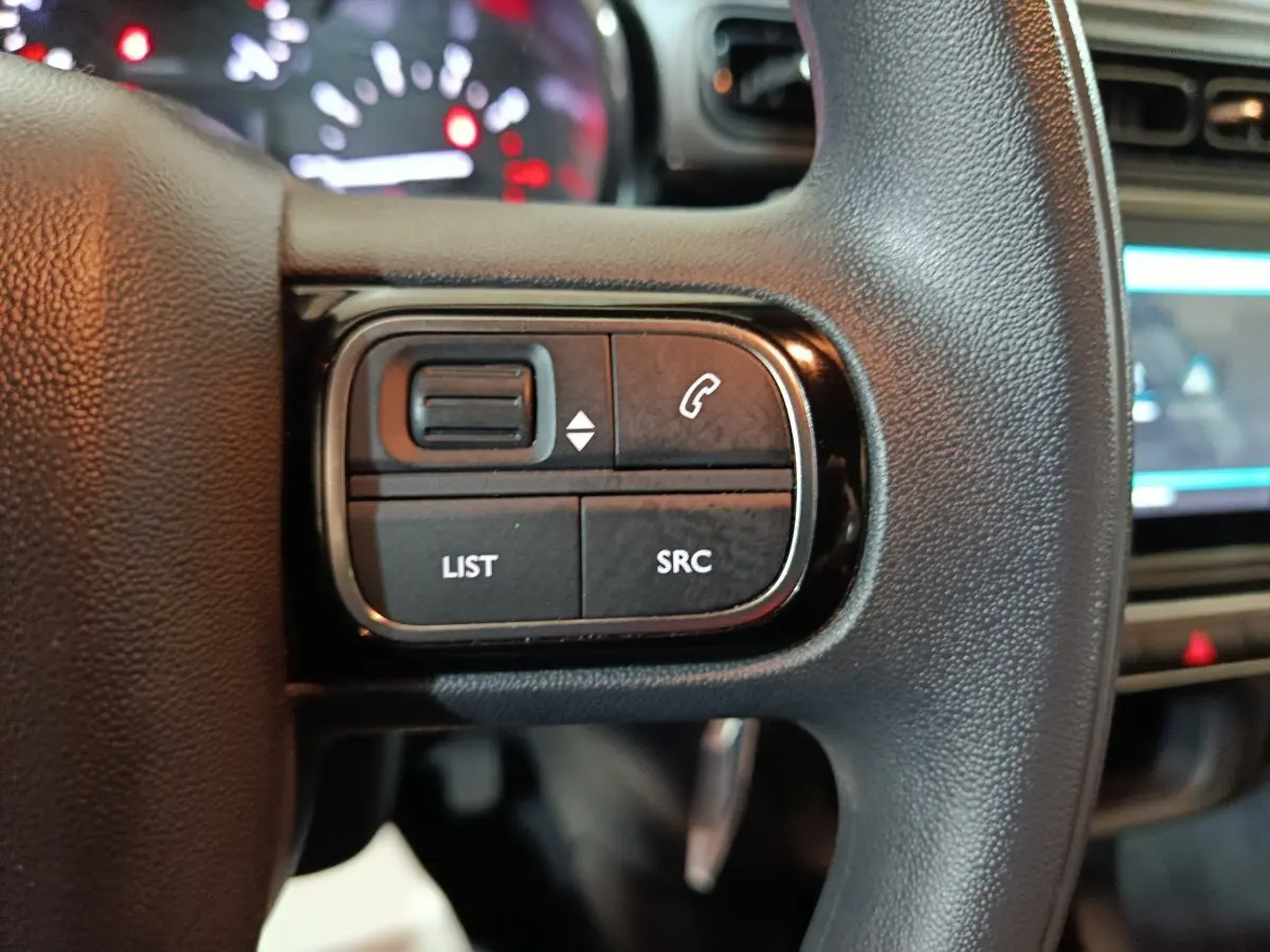 Gros plan sur les commandes gauche du volant noir d’une Citroën C3 2024, avec tableau de bord et écran tactile en arrière-plan.
