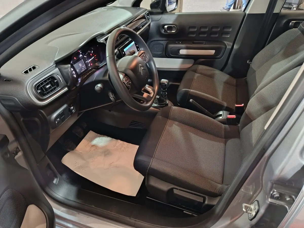 Vue intérieure côté conducteur d'une Citroën C3 gris clair 2024, montrant le tableau de bord et les sièges tissu noirs.