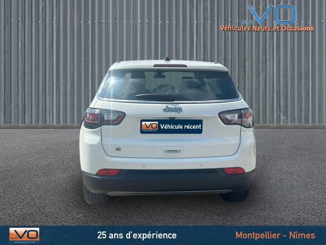 Vue arrière d'un Jeep Compass blanc 2024 avec logo et feux sombres, stationné devant un mur métallique.