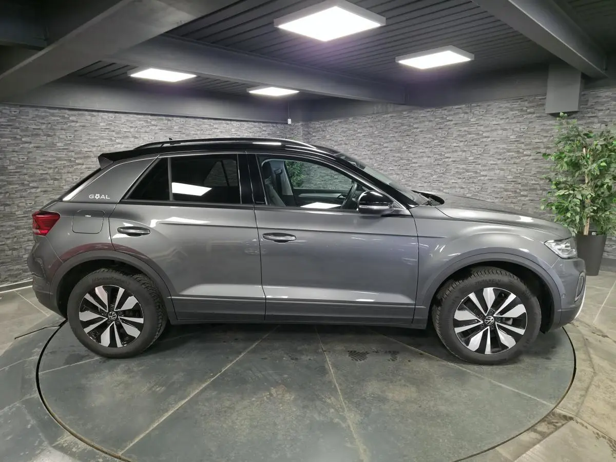 Vue latérale droite d'un Volkswagen T-Roc gris métallisé édition GOAL avec toit et montants noirs en intérieur showroom.