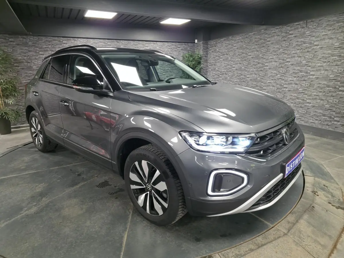 Volkswagen T-Roc gris 3/4 avant droit en intérieur, avec phares LED allumés et jantes alliage 17 pouces.