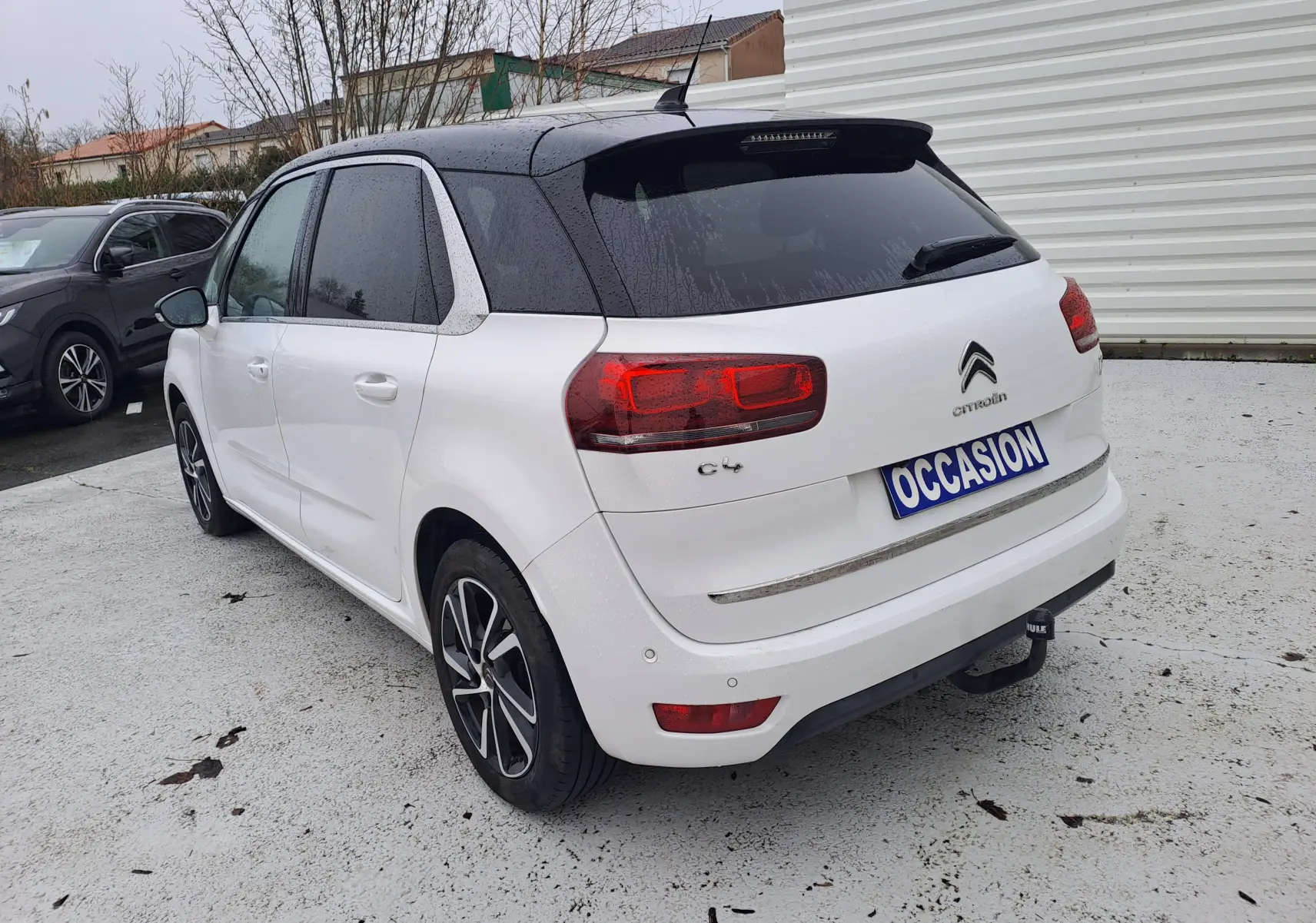Vue 3/4 arrière droite d'une Citroën C4 Spacetourer blanche avec toit noir et jantes bi-ton, sur parking extérieur humide.