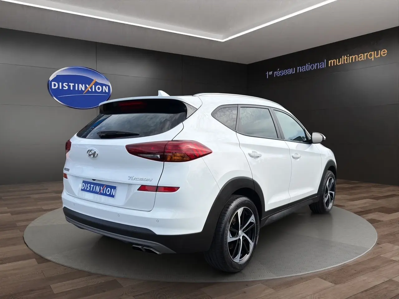 Vue 3/4 arrière droite d'un Hyundai Tucson blanc polar avec jantes 18 pouces bicolores, sur plateforme en showroom.