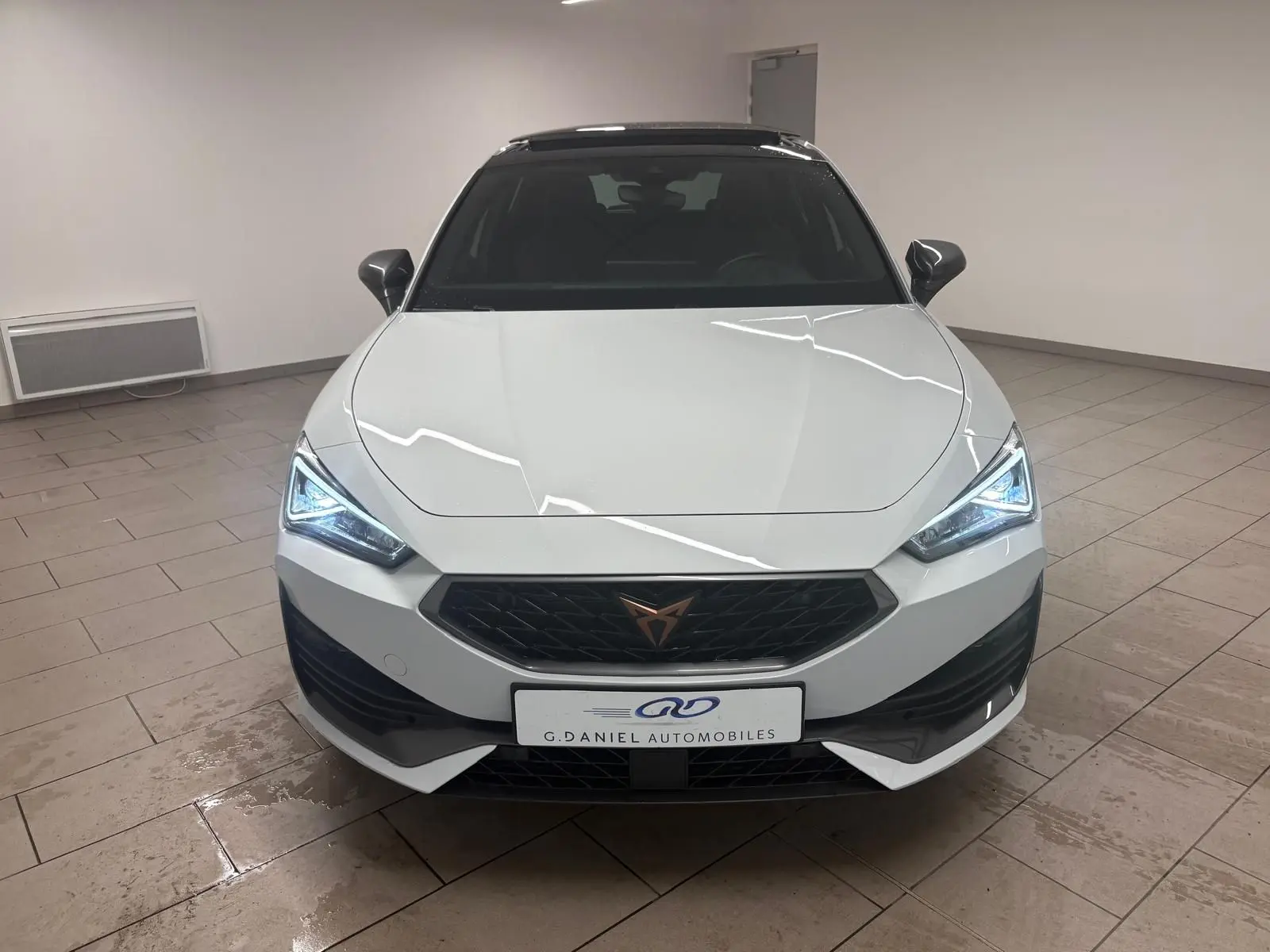 Vue frontale d'une CUPRA Leon blanche Nevada avec toit ouvrant panoramique et phares LED allumés en intérieur.