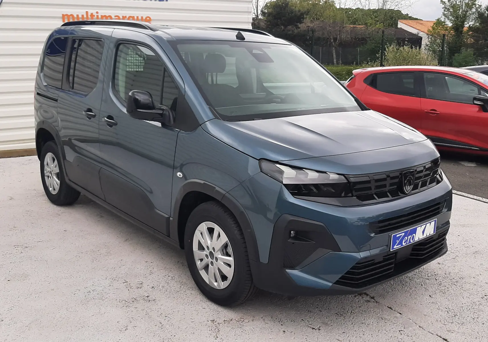 Peugeot Rifter bleu Kiama vu en 3/4 avant droit, avec calandre noire et jantes argentées, stationné devant un mur blanc.