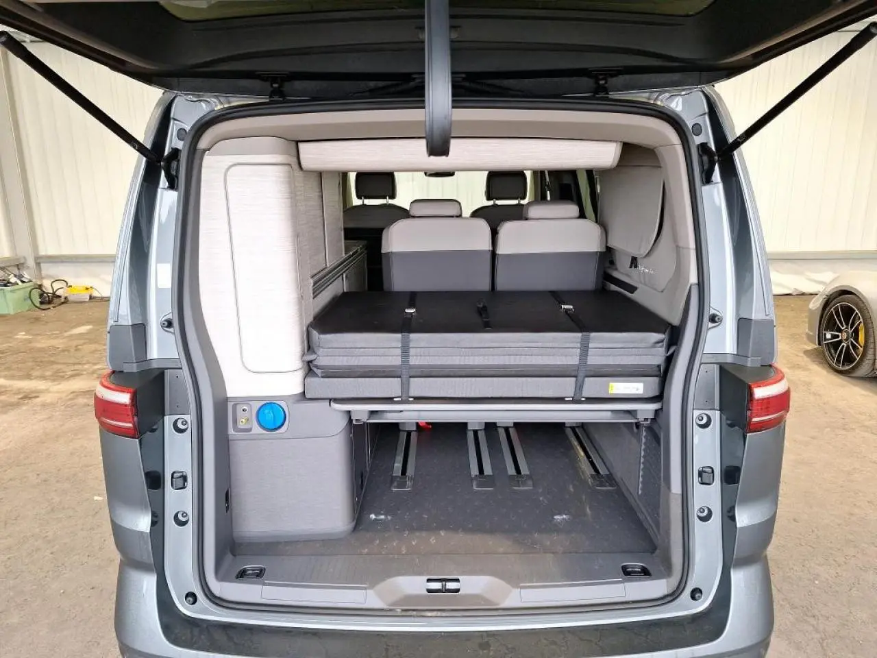 Vue arrière du Volkswagen California T7 gris Indium avec hayon ouvert montrant l'intérieur modulable et la couchette repliée.