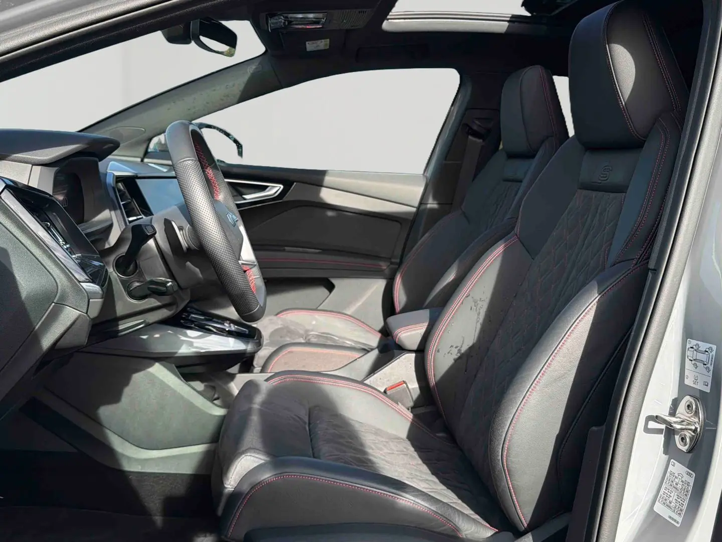 Vue intérieure côté gauche de l'Audi Q4 e-tron 2025, sièges en cuir noir avec surpiqûres rouges et volant multifonction.
