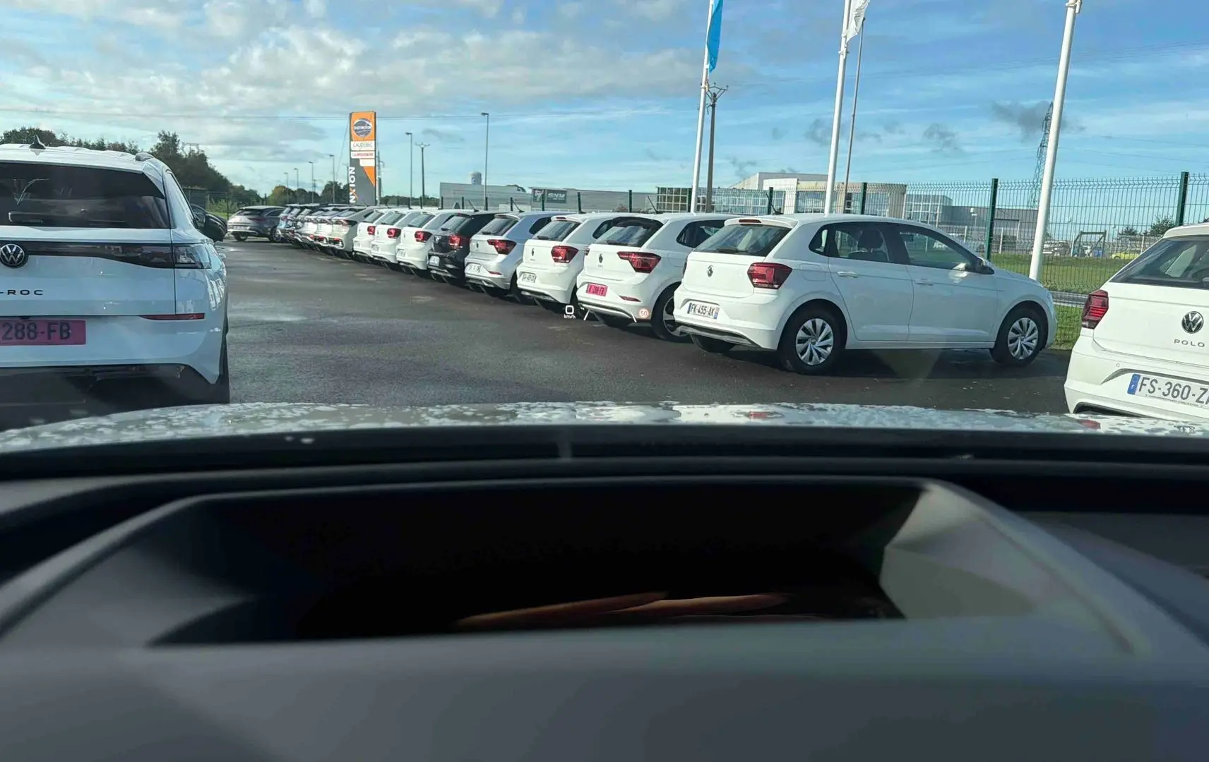 Vue depuis l'intérieur montrant le tableau de bord et plusieurs Volkswagen blanches garées à l'extérieur.