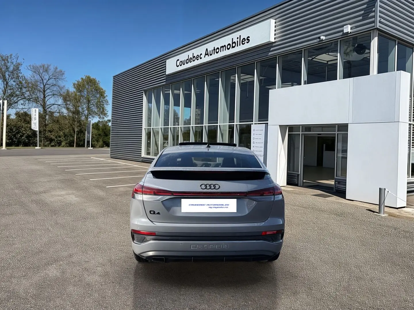 Vue arrière d'un Audi Q4 e-tron gris galet stationné devant un bâtiment moderne de concession automobile.