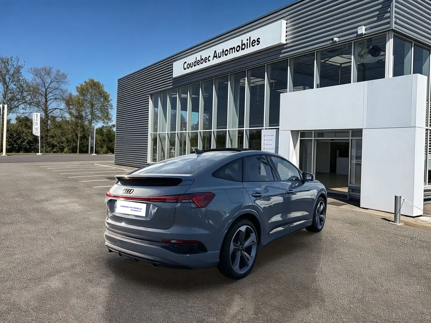 Audi Q4 e-tron gris galet en 3/4 arrière droit, avec jantes 19 pouces, stationné devant un bâtiment moderne.