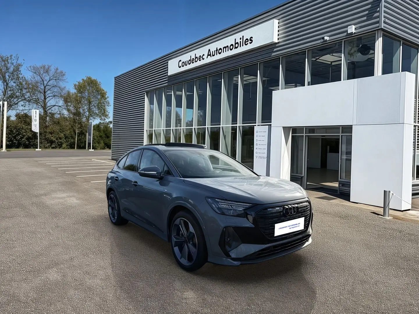 Audi Q4 e-tron gris galet en 3/4 avant droit devant un bâtiment de concession Coudebec Automobiles.