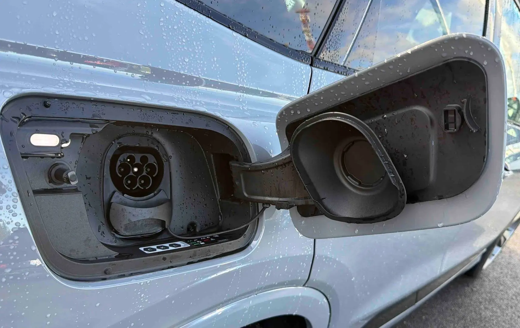 Prise de recharge électrique ouverte côté gauche sur Audi Q4 e-tron gris galet avec gouttes d'eau visibles.