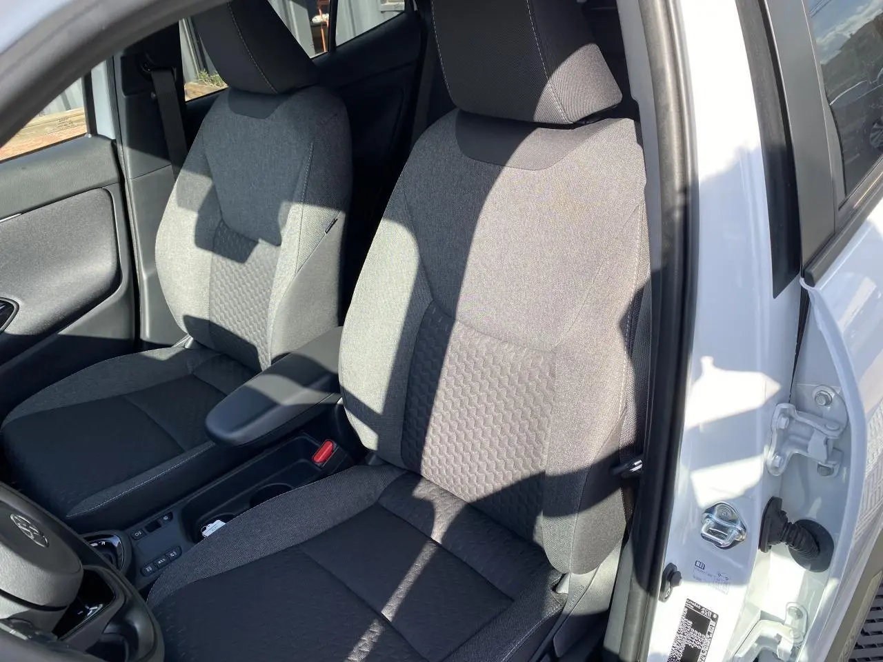 Vue rapprochée des sièges avant gris tissu du Toyota Yaris Cross blanc, avec accoudoir central et portière ouverte côté gauche.