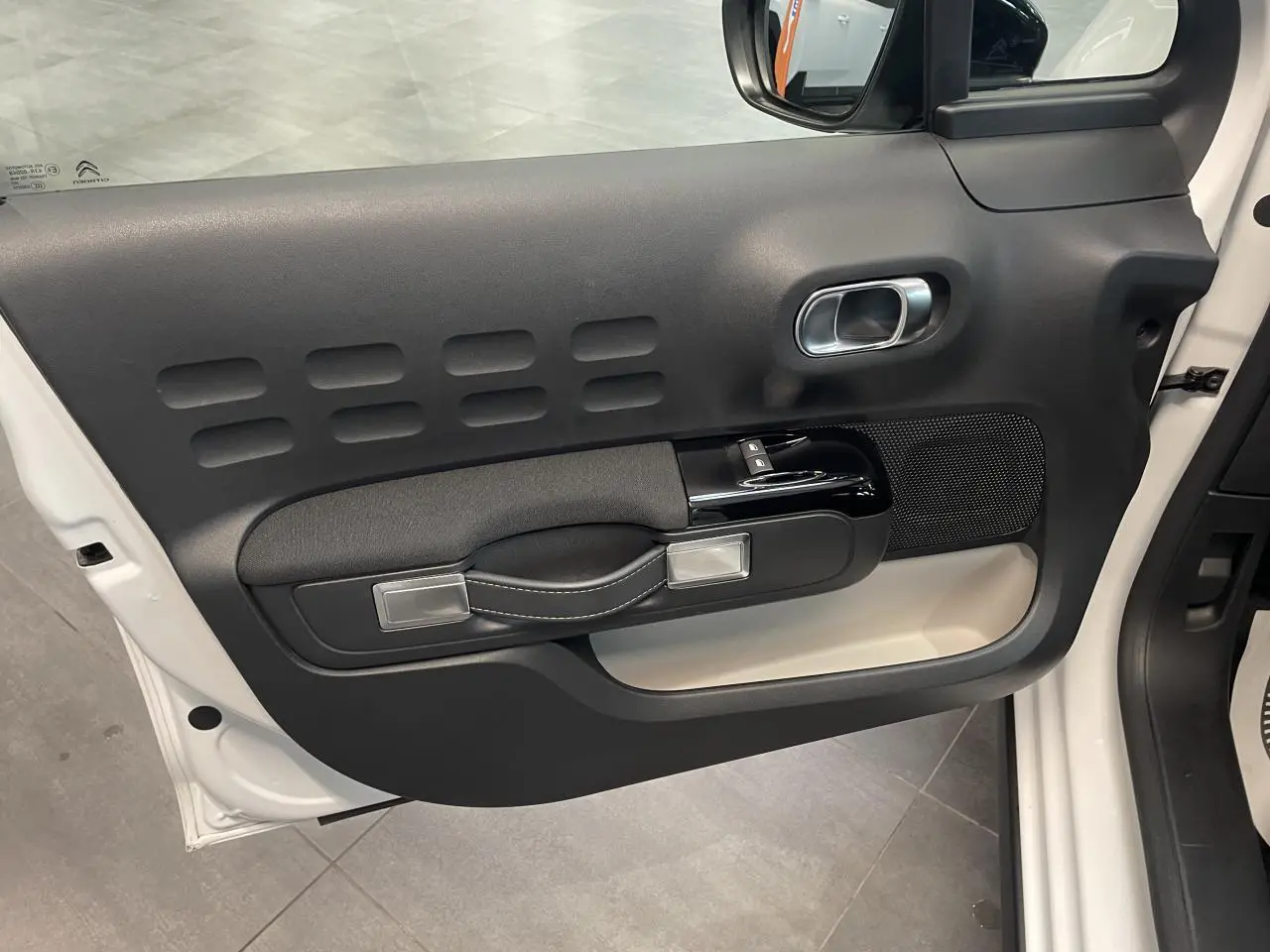 Vue intérieure de la porte avant gauche blanche de la Citroën C3 2023 avec poignée textile et commandes électriques.
