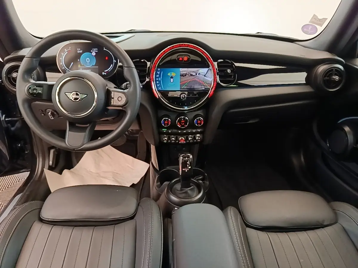 Vue intérieure avant de la MINI Cooper Premium Plus 2023, tableau de bord noir avec écran central rond et volant cuir multifonctions.