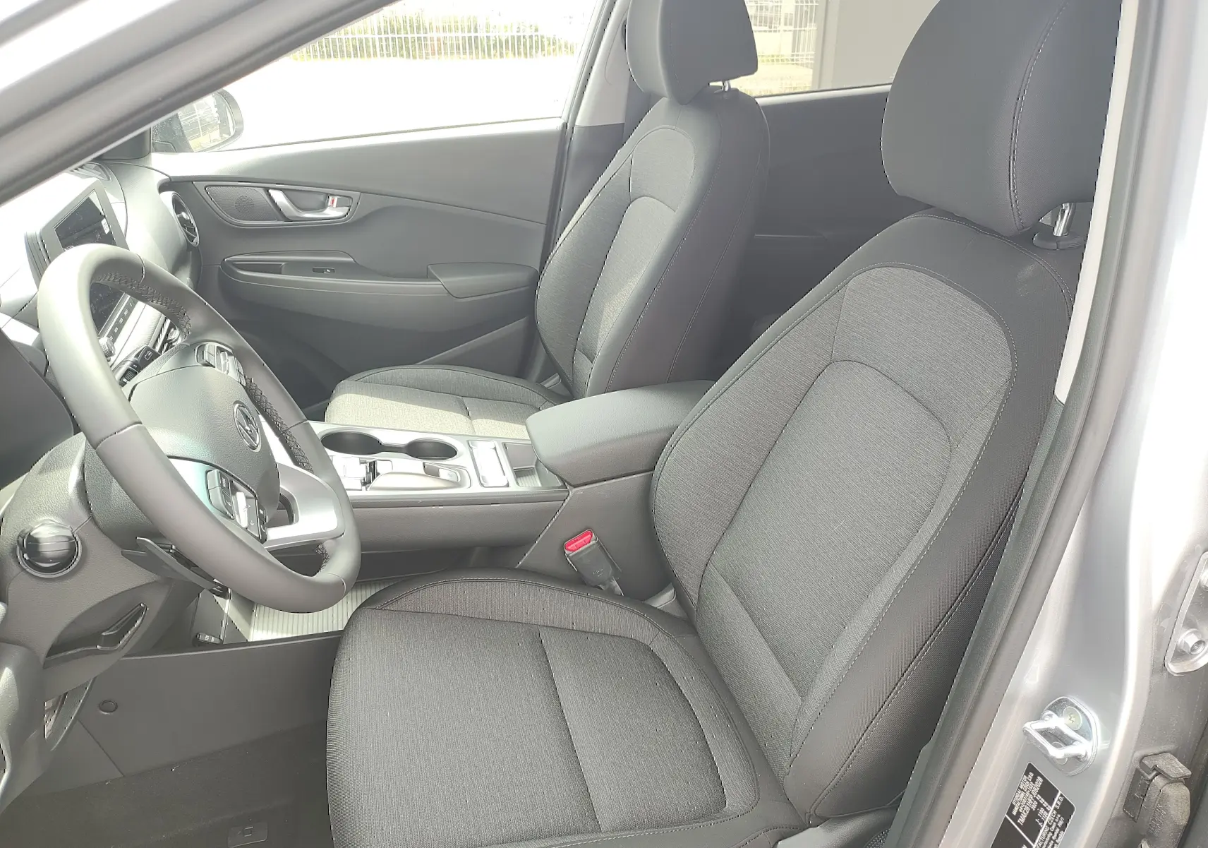 Vue intérieure côté conducteur du Hyundai Kona Electric gris, sièges tissu noir et console centrale avec accoudoir.