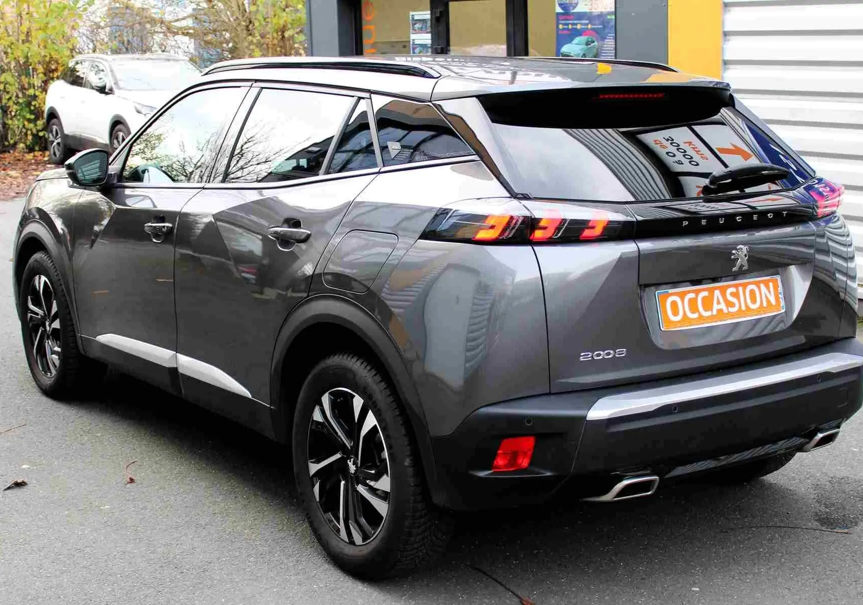 Peugeot 2008 gris platinium vue 3/4 arrière droit, feux arrière allumés et jantes noires bicolores.