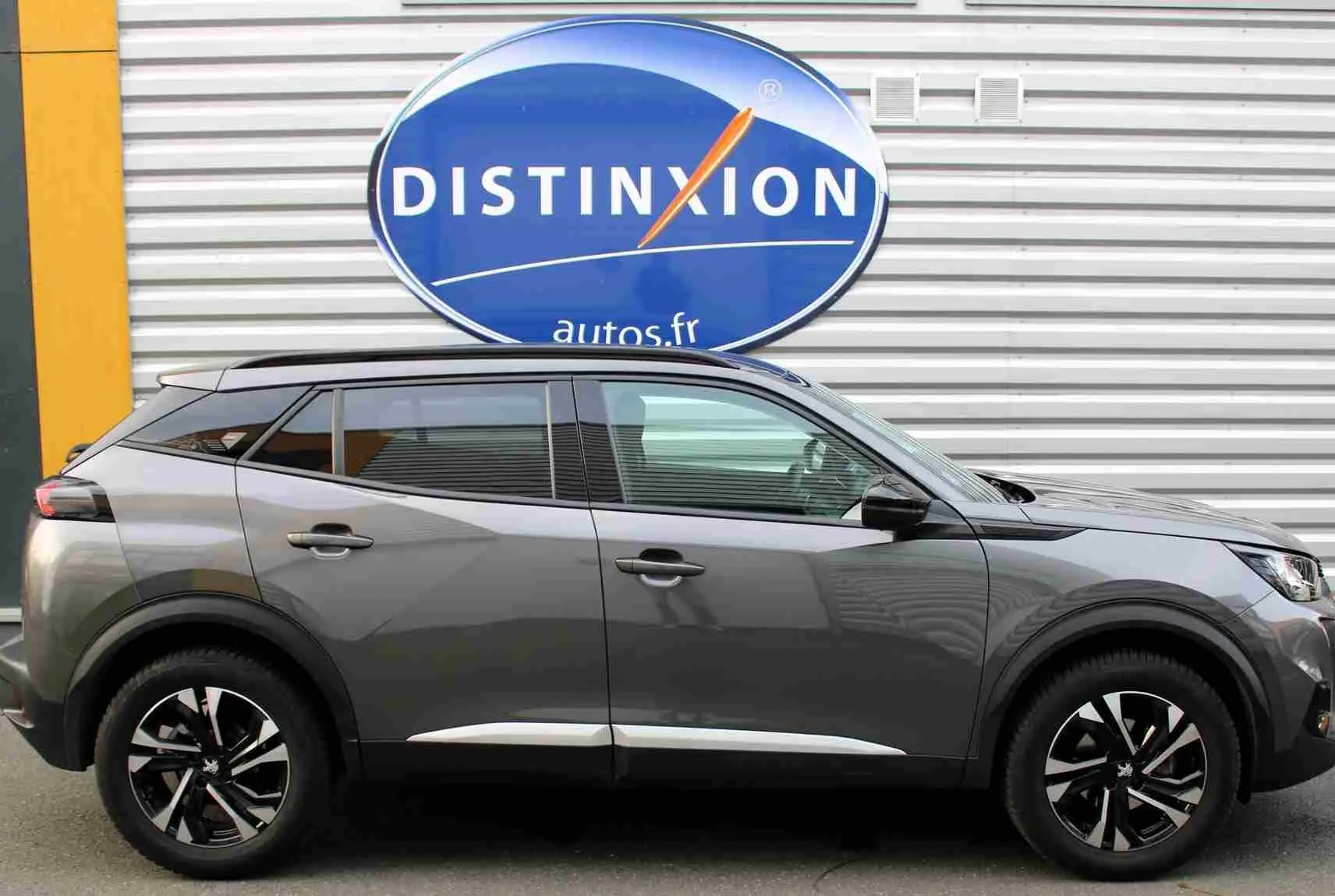 Profil droit d'un Peugeot 2008 gris platinium avec jantes noires et toit panoramique visible, devant un mur métallique.