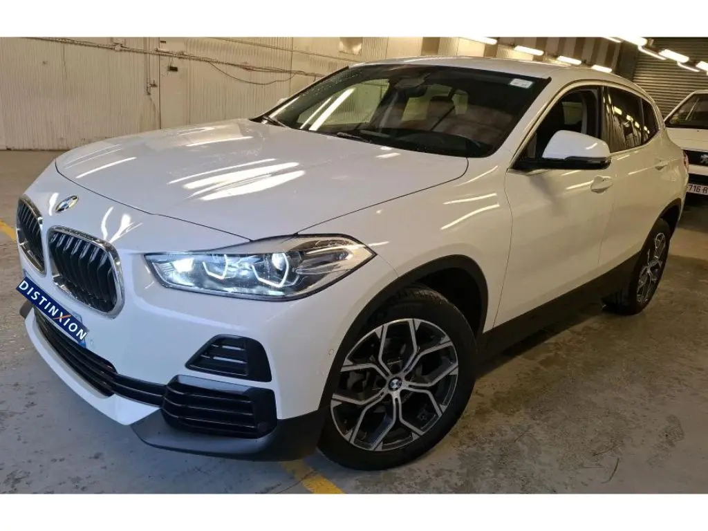 BMW X2 blanc vue 3/4 avant droit, jantes alliage 18'', rétroviseurs électrochromes et phares allumés.