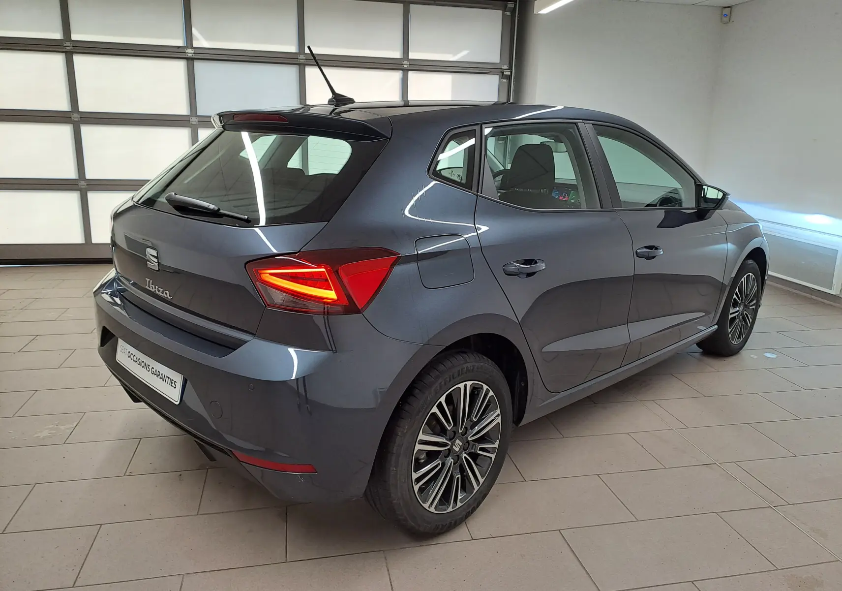 Vue 3/4 arrière droite d'une SEAT Ibiza 2025 gris magnétique avec feux arrière allumés dans un garage.