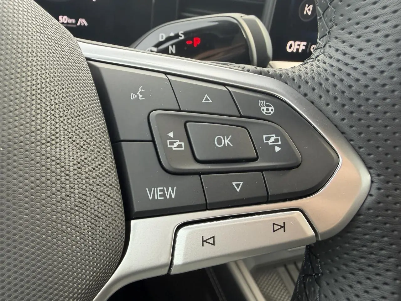 Gros plan sur les commandes noires et argent du volant perforé du Volkswagen T-Roc 2026, avec affichage numérique en arrière-plan.