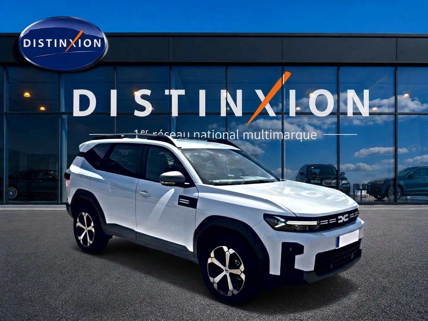 Vue 3/4 avant droit du SUV blanc Dacia Bigster 2026 avec barres de toit et jantes alu distinctives, devant un showroom Distinxion.